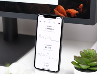 A smartphone displays analytics data with graphs showing views, downloads, and likes. It is resting vertically on a white dock next to a small potted succulent plant. In the background, a large computer monitor is partially visible, along with a closed laptop and a white wireless earbud case.