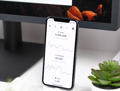 A smartphone displays analytics data with graphs showing views, downloads, and likes. It is resting vertically on a white dock next to a small potted succulent plant. In the background, a large computer monitor is partially visible, along with a closed laptop and a white wireless earbud case.