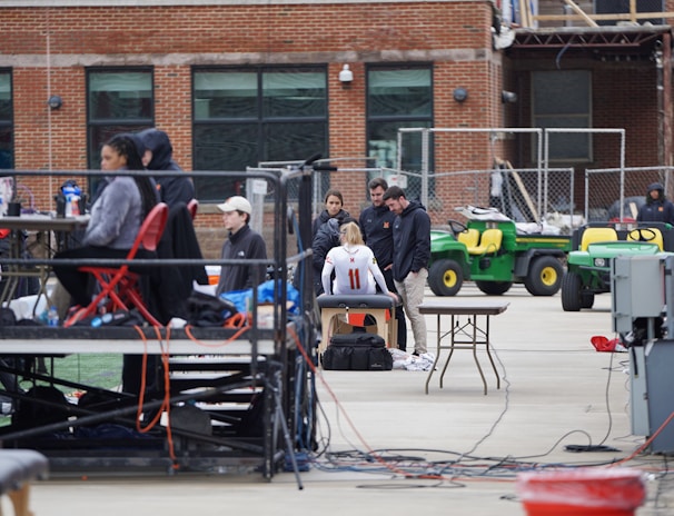 A group of people are gathered around an individual wearing a sports jersey with the number 11. They appear to be in a sports facility area, possibly a training or medical setup. There are tables, chairs, and equipment scattered around, with a brick building in the background. Some individuals are seated on a platform with electronics close by, suggesting a recording or monitoring activity. Utility vehicles and an orange barrier are present in the background.