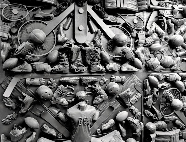 A complex arrangement of various sports equipment and attire, including tennis rackets, soccer balls, basketballs, helmets, shoes, gloves, and a mannequin torso wearing a T-shirt. The items are layered and artistically displayed in a monochrome color scheme, creating an intricate texture and visual pattern.
