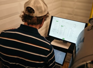A person with a striped shirt and a cap is standing in front of a computer screen displaying data and analytics. Another laptop is open on a desk below the monitor, and a large metallic tank with a pressure gauge is nearby.
