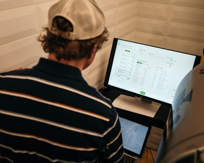 A person with a striped shirt and a cap is standing in front of a computer screen displaying data and analytics. Another laptop is open on a desk below the monitor, and a large metallic tank with a pressure gauge is nearby.
