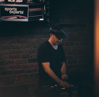 A person wearing a hat is focused on operating DJ equipment in a dimly lit room with a brick wall. Above this setup, a television screen displays a broadcast featuring three individuals discussing sports.