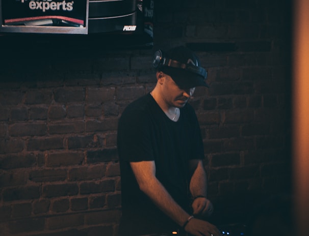 A person wearing a hat is focused on operating DJ equipment in a dimly lit room with a brick wall. Above this setup, a television screen displays a broadcast featuring three individuals discussing sports.