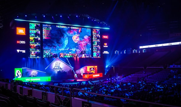 A large esports arena featuring a massive screen displaying a video game, surrounded by sponsor logos and team names. The auditorium is filled with spectators watching the gaming event. Neon blue and purple lights illuminate the stage, creating a vibrant atmosphere.