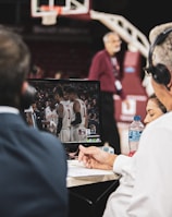 Two men wearing headsets focus on a monitor displaying a basketball team in discussion. Others can be seen around them, including a woman with a bottle of water, and an ongoing basketball game in the background.