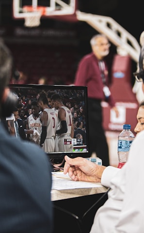 Two men wearing headsets focus on a monitor displaying a basketball team in discussion. Others can be seen around them, including a woman with a bottle of water, and an ongoing basketball game in the background.