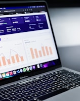 A laptop with an open screen displaying dashboard analytics featuring charts, graphs, and numerical data related to sales and subscriptions. The interface is predominantly dark-themed with sections highlighting key performance metrics. The device shown is a MacBook Pro, indicated by the keyboard layout and branding.