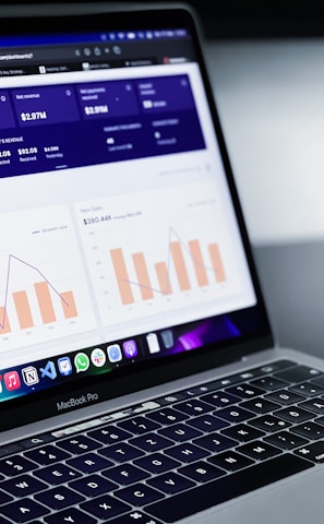 A laptop with an open screen displaying dashboard analytics featuring charts, graphs, and numerical data related to sales and subscriptions. The interface is predominantly dark-themed with sections highlighting key performance metrics. The device shown is a MacBook Pro, indicated by the keyboard layout and branding.