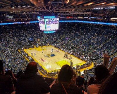 An indoor basketball arena filled with spectators. The court features a central logo and is surrounded by partially occupied tiered seating. A large scoreboard hangs above the center of the court, displaying the game score and time. Ambient lighting highlights the players on the court and the vibrant atmosphere of a live sports event.