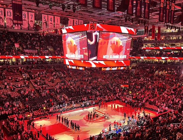An indoor sports arena filled with spectators and a basketball court illuminated by red lighting. The scoreboards and video screens display promotional content, with numerous banners and championship flags hanging from the ceiling. Players and officials are gathered on the court, possibly in preparation for a game or event.