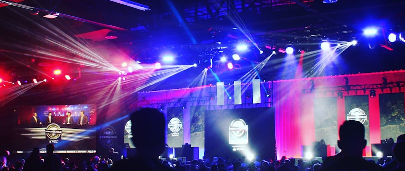 A dimly lit indoor arena filled with an audience. Bright stage lights in red and blue illuminate multiple large screens displaying graphics.
