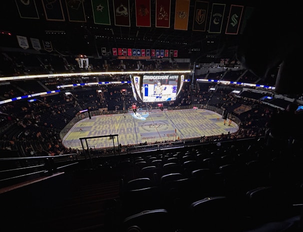 An indoor ice hockey arena with an illuminated scoreboard and a partially filled audience. The ice rink is visible with graphics projected onto the surface, and banners hang from the ceiling. Players are on the ice, and spectators are scattered throughout the stands.