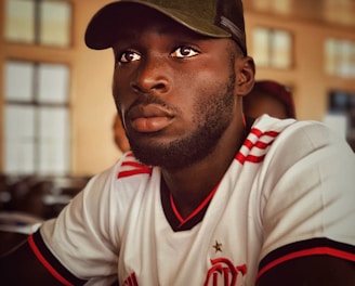 A person is wearing a white and red sports jersey with logos and is seated indoors. They have a short beard and are wearing a dark green cap. The background features a softly lit room with blurred windows and ceiling fans.