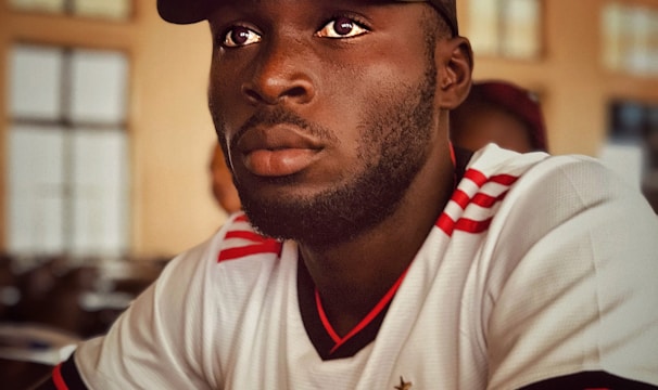 A person is wearing a white and red sports jersey with logos and is seated indoors. They have a short beard and are wearing a dark green cap. The background features a softly lit room with blurred windows and ceiling fans.